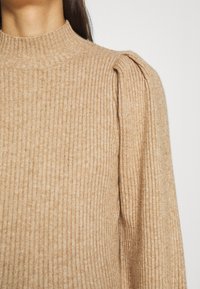 Beiger, gerippter Pullover mit hohem Kragen und Bubikragenärmeln, mit weicher Textur und subtiler Mischung aus hellen und dunklen Fäden.