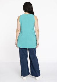 Ein ärmelloses türkisfarbenes Oberteil mit rundem Ausschnitt, kombiniert mit weiten dunkelblauen Jeans und weißen Turnschuhen, von hinten betrachtet.