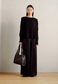 Maglietta nera a maniche lunghe abbinata a una gonna maxi nera fluente. Una borsa di pelle nera con manico circolare completa l'outfit, su uno sfondo neutro.