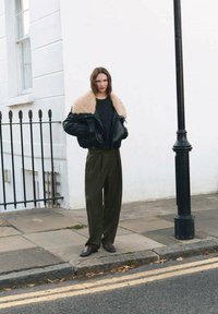 Chaqueta de cuero negra con un cuello de borrego beige, combinada con pantalones de pierna ancha en verde oliva y zapatos oscuros, con un fondo de pared blanca.