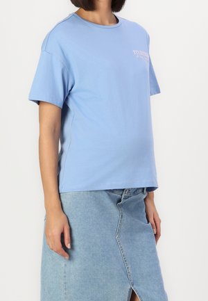 Kobieta ubrana w jasnoniebieską koszulkę z nadrukiem oraz jasnoniebieską spódnicę jeansową z przednim rozcięciem, stojąca na tle gładkiej białej ściany.