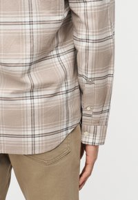 Μανσέτα και πίσω μέρος πουκάμισου μακρυμάνικου με καρό μπεζ και λευκό, με τσέπη από ανοιχτό καφέ παντελόνι και ένα χέρι ορατό στο πλάι.