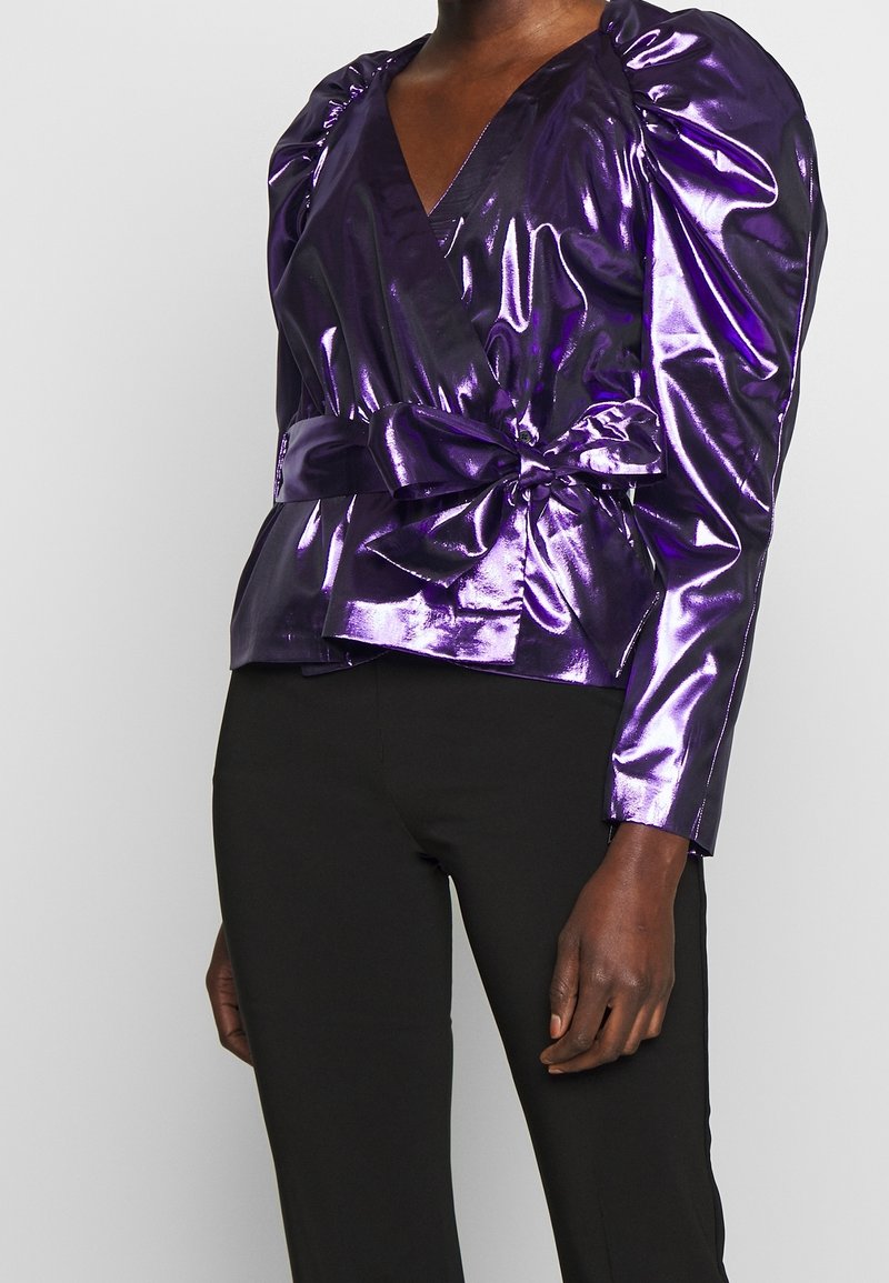 Blouse en tissu métallique violet avec un décolleté en V profond, des manches bouffantes et une taille nouée. Le tissu a une texture brillante et réfléchissante. Associée à un pantalon noir.