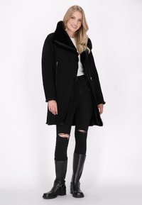 Cappotto in pelliccia sintetica nera foderato con ampio collo, tasche con zip e taglio dritto. Abbinato a jeans skinny neri e stivali alti fino al ginocchio.