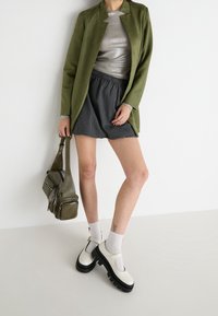 Chaqueta larga verde oliva, top plateado metálico, pantalones cortos a rayas grises, zapatos de plataforma blancos y bolso verde con múltiples bolsillos.