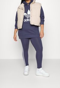 Beige pufferväst över en marinblå hoodie med vit logotyp, kombinerad med marinblå leggings med vita sidostripes och vita sneakers.