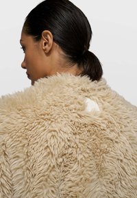 Abrigo de piel sintética beige con una superficie texturizada y esponjosa. Presenta una pequeña etiqueta blanca en la parte trasera, con cuello redondo y caída suave.