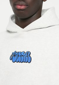 Sudadera gris claro con un gráfico bordado en azul, con letras negritas y redondeadas contorneadas en negro, cerca del cuello.