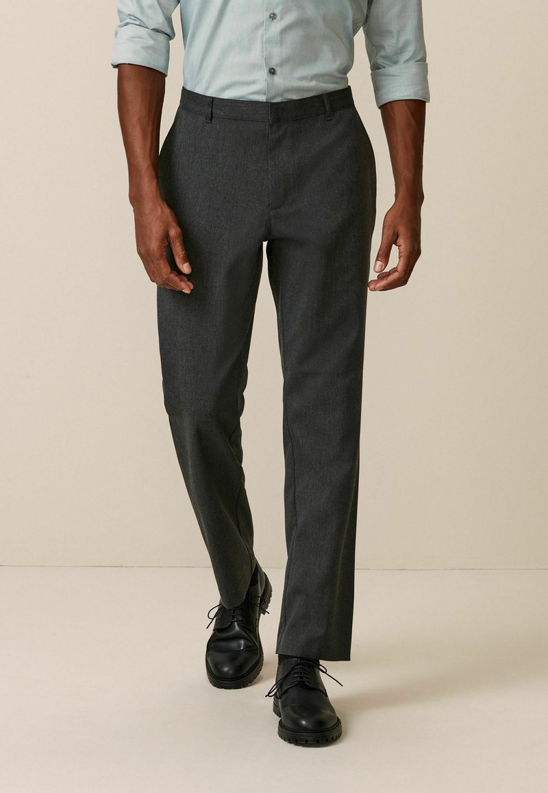 Un pantalon gris foncé, ajusté, avec une texture lisse et une coupe droite. Porté avec une chemise à boutons bleu clair et des chaussures noires.