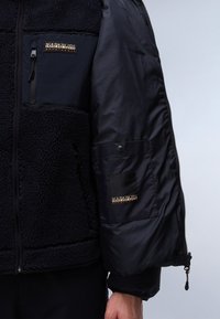 Fleece-fütterte Jacke mit schwarzem Nylon-Außenmaterial, Reißverschlusstaschen und Logo-Akzenten auf der Brust und dem Ärmel. Verfügt über ein Pufferdesign.