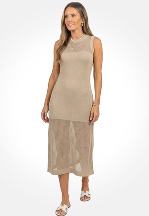 Vrouw met lang haar, gekleed in een mouwloze beige gebreide midi-jurk met inzetstukken van mesh, witte sandalen en gouden sieraden, staand tegen een witte achtergrond.