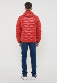 Giacca puffer rossa con cappuccio, caratterizzata da un design trapuntato e una finitura lucida. Indossata con jeans blu e sneaker nere, vista da dietro.