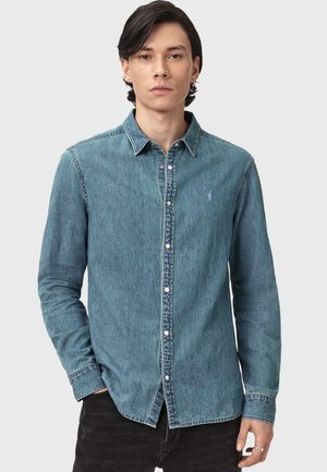 Mann trägt ein langärmeliges, blaues Jeanshemd mit Knöpfen und Kragen, steht vor einem schlichten hellen Hintergrund.