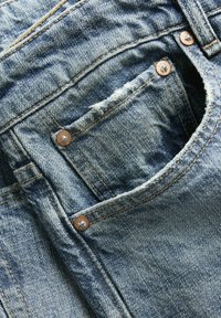 Nahaufnahme einer blauen Jeans-Tasche mit sichtbaren Nähten, Kupfernieten und leicht ausgefranstem Rand an der kleinen Münztasche.