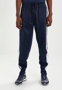 Marinblåa träningsbyxor gjorda av glatt tyg, med vita sidsnoddar och ett elastiskt midjeband med dragsko för justerbar passform.