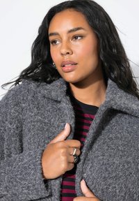 Cappotto grigio testurizzato con colletto a lancia, con chiusura frontale a bottoni. Il modello indossa una camicia a righe sotto, mostrando un look informale.