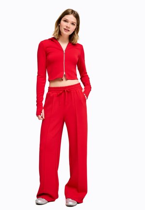 Ung kvinde iført en kort rød lynlåstrøje og vide røde bukser med sølvfarvede sneakers, stående med den ene hånd i lommen.