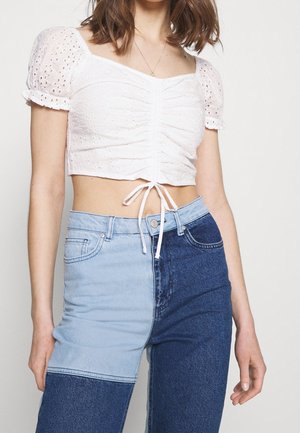 Kobieta w białym krótkim topie z ażurowymi bufkami, marszczonym wiązaniem z przodu oraz dwukolorowych jeansach z wysokim stanem w jasnym i ciemnym denimu.