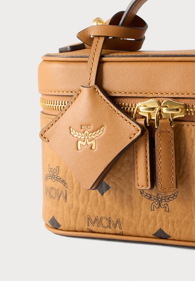 Sac en cuir fauve avec une finition texturée, fermetures éclair dorées, étiquette en forme de losange ornée du logo et accents de motifs sombres.