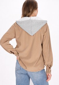 Cămașă din corduroy maro cu hanorac gri, având o croială dreaptă, manșete cu nasturi și textură verticală în relief.