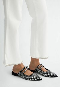 Schwarze spitze Ballerinas mit Strassdetails, aus glattem Material und zwei Knöchelriemen. Kombiniert mit weißen, ausgestellt geschnittenen Hosen mit Fransen.