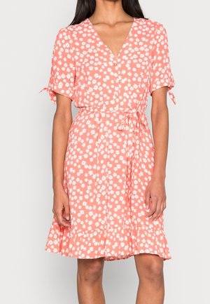 Femme portant une robe portefeuille corail avec un motif floral blanc, manches courtes nouées aux extrémités et un ourlet volanté tombant au-dessus des genoux.