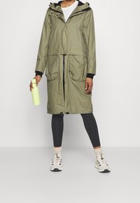 Oliwkowy wodoodporny płaszcz z kapturem, zamek błyskawiczny z przodu, dwie duże kieszenie i czarne mankiety, noszony na czarnych legginsach i białych sneakersach.