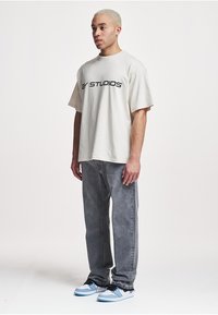 Oversized cream T-shirt with "2Y STUDIOS" printed in black, paired with gray jeans and blue-and-white sneakers. Smooth fabric and relaxed fit.
