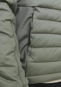 Padded Jacke in Staubgrün, mit glattem Außenmaterial, gesteppten Abschnitten und einer seitlichen Reißverschlusstasche für sicheren Stauraum.