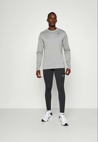 Chemise grise à manches longues avec un logo noir sur la poitrine ; leggings noirs ajustés ; baskets blanches avec accents noirs ; se tenant devant un fond neutre.