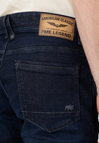 Tamno plave traperice od denima, s smešnim kožnim obeležjem s utisnutim tekstom i logotipom na zadnjem pojasu, te suptilnim detaljima šivanja.