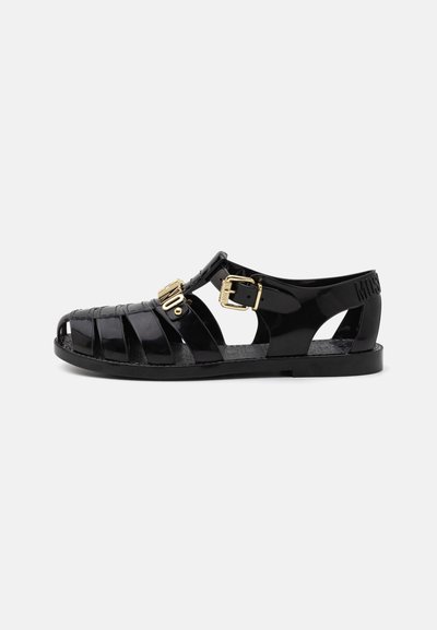 Zwarte plastic sandalen met een riemontwerp, gesloten neus en gouden gespen. De zool is vlak met textuurelementen en branding.