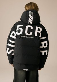 Personne portant une veste rembourrée noire avec le texte en gras "SUB5CRIBE" en blanc et des coordonnées imprimées au dos et sur les manches.