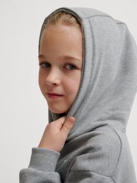 Lille barn med blå øjne trækker hætten på en grå sweatshirt op og kigger let til siden mod en ensfarvet baggrund.