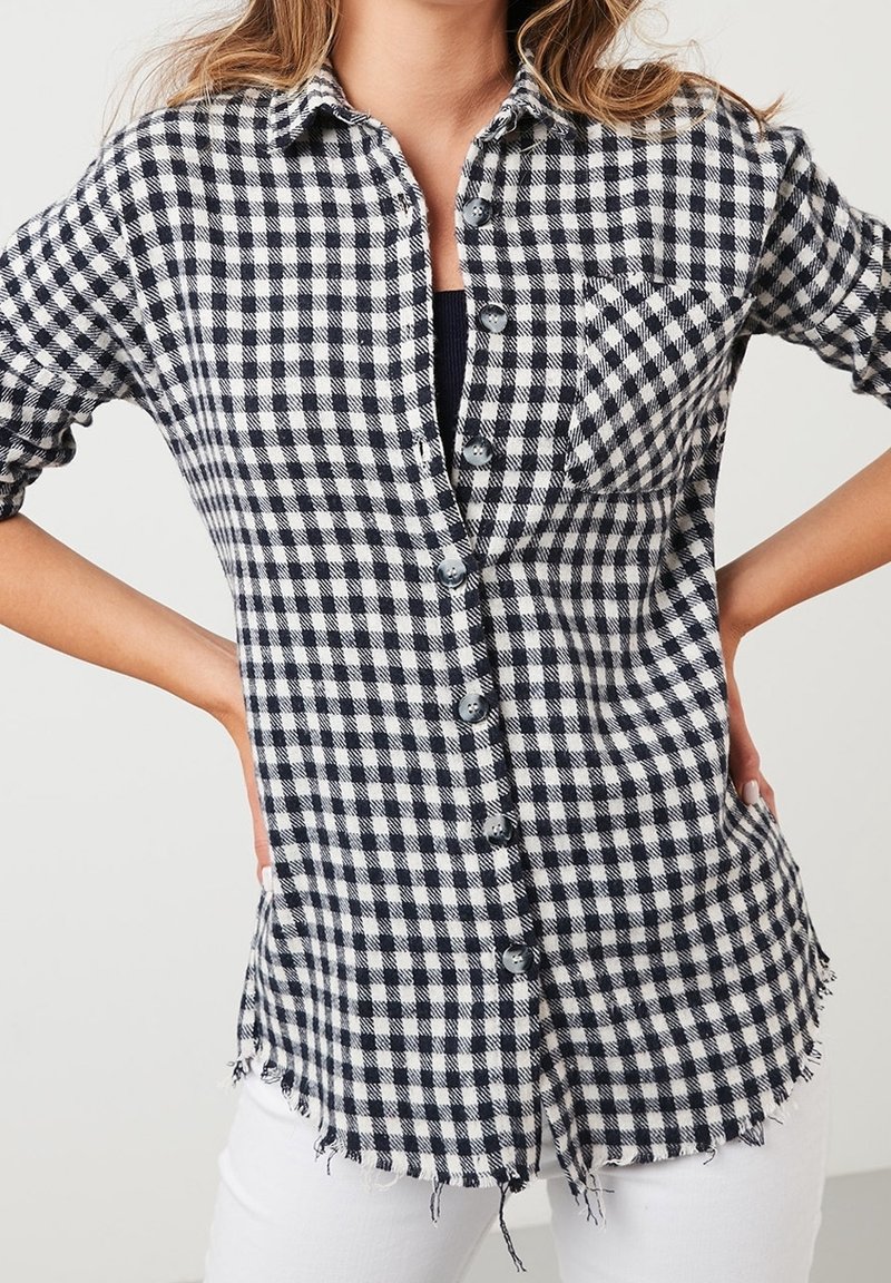 Femme portant une chemise à carreaux noir et blanc avec un ourlet effiloché et un pantalon blanc, debout, les mains sur les hanches.