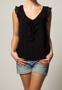 Svart blus med V-ringning och volangdetaljer vid axlarna och fram, tillsammans med ljusblå uppvikta denimshorts.