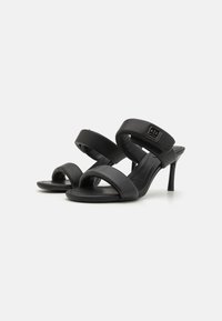 Zwarte hoge hakken sandalen met twee brede banden, gemaakt van soepel faux leer, met een logo-accent op de bovenste band.