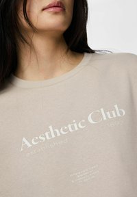 Moteris dėvi smėlio spalvos marškinius, ant kurių priekyje parašyta „Aesthetic Club įsteigtas 1892“ ir mažesnis šūkis apie stilių, išraišką ir kūrybiškumą.