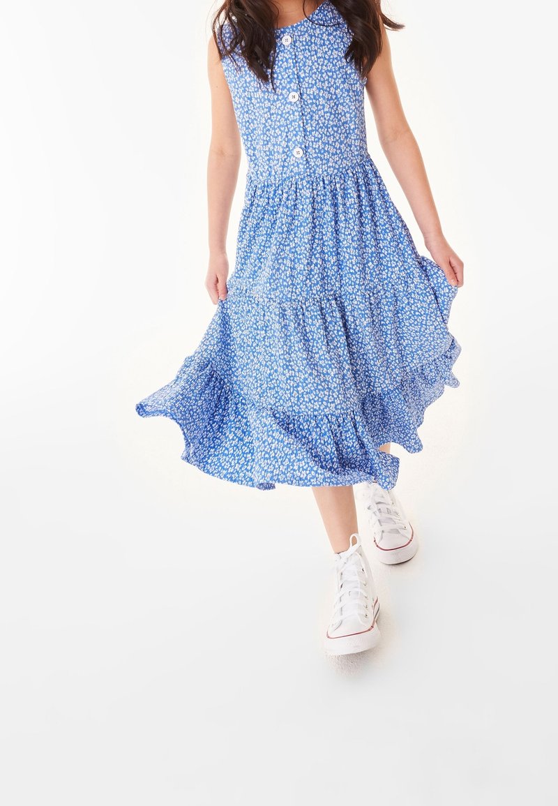 Fille portant une robe bleue fleurie sans manches avec des boutons et des baskets blanches, tenant sa robe en marchant sur un fond blanc.