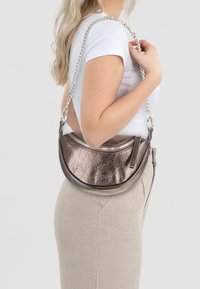 Borsa in bronzo metallico con forma curvilinea, chiusura con zip e tracolla a catena. La superficie presenta una texture rugosa, riflettendo la luce.