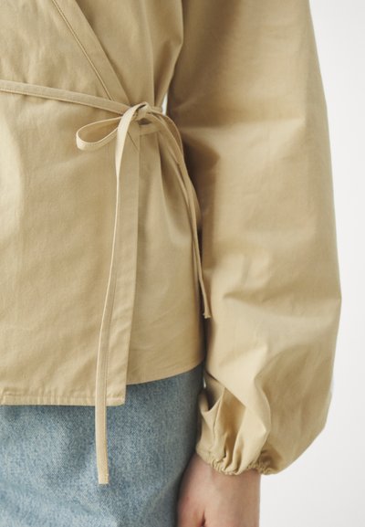 Beige wikkeltop met lange, puffy mouwen en een gestrikte taille. Gemaakt van soepele stof, met een strak ontwerp en een relaxte pasvorm.