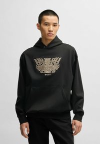 Svart hoodie med ett stort, intrikat guldemblem i mitten. Materialet verkar slätt och har en framficka med kangaroo-design.