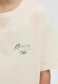 Kurzärmliges T-Shirt in Creme mit Rundhalsausschnitt, bedruckt mit den Textgrafiken "Casares" und "Malaga" sowie einem kleinen Etikett mit der Aufschrift "Das kreative Studio."