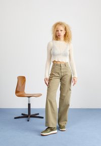 Ljusbeige stickad crop top med vågiga kanter, ihop med olivgröna cargobyxor. Modellen bär gröna och bruna sneakers. Trästol i bakgrunden.