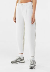 Pantalones de mezclilla blancos con corte recto, que presentan paneles laterales beige claro y un cierre de botón clásico. Combinados con zapatillas grises.