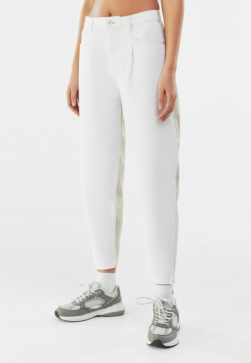 Pantalones de mezclilla blancos con corte recto, que presentan paneles laterales beige claro y un cierre de botón clásico. Combinados con zapatillas grises.