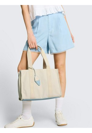 Persoon draagt lichtblauwe korte broek en witte sneakers, en houdt een beige totebag vast met "STAY COOL" bandjes en een ijsjessluier.