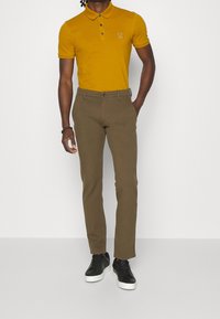 Man in een mosterdgele short-sleeve polo, olijfgroene rechte broek, zwarte sneakers met witte zolen en een zwarte armband.