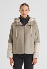 Beige Zip-Jacke mit einem strukturierten Wollkörper und glänzenden, puffigen Ärmeln. Mit Kapuze und Kordeln, getragen über einem dunklen Rollkragenpullover.