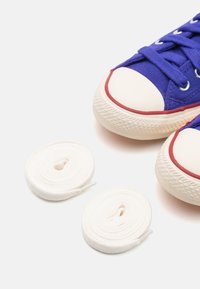 Baskets en toile bleue avec embouts en caoutchouc blanc et accents rouges. Deux lacets blancs roulés placés à côté des chaussures. Détails de la semelle texturée visibles.